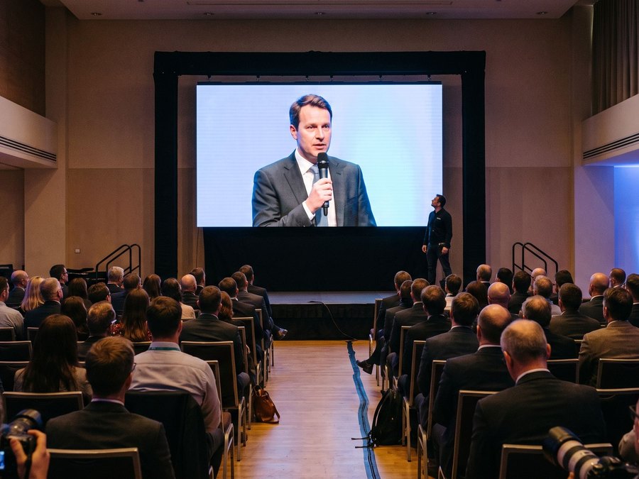 LED-wand op het podium van een zaal met publiek, een spreker in beeld